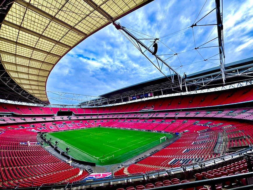 Interior view of the empty, modern stadium with multiple tiers of red seating, large overhead screen, and glass facades letting in natural light. The central pitch is well-maintained with lush green g